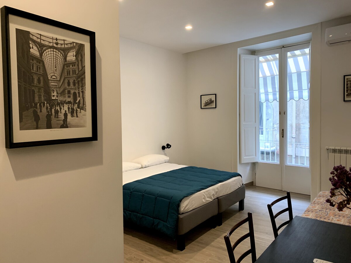A bright and airy room is presented, featuring a spacious bed with a blue quilt. Natural light enters through a large window adorned with striped curtains. A framed black-and-white photograph hangs on the wall, and a small dining area is visible with a dark table and chairs.