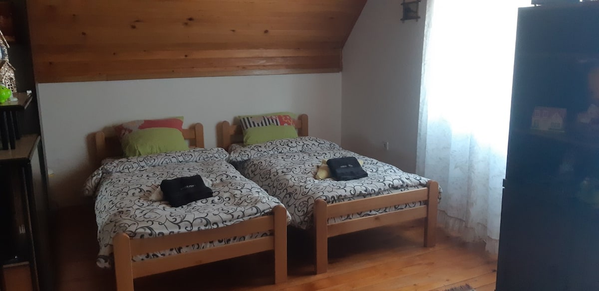 Two separate beds are placed in a room with wooden accents, featuring light wood frames and decorative bedding. Natural light enters through a window draped with sheer curtains, illuminating the space. A dark furniture unit stands in one corner, contributing to the room's cozy atmosphere.