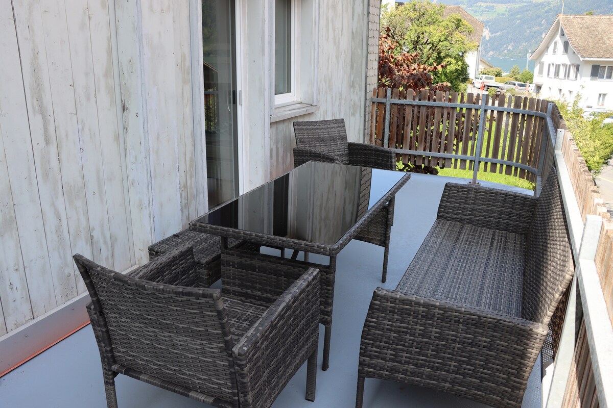 A patio area features a black glass-topped table surrounded by four woven rattan chairs. The space is enclosed by wooden railings and offers views of the surrounding landscape, providing a serene spot for outdoor dining or relaxation.