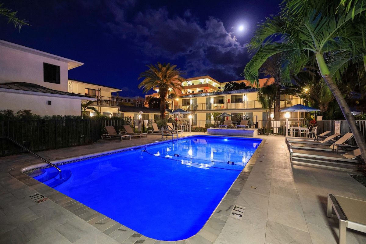 An inviting pool area is illuminated under the moonlight, featuring a clear blue pool with lounge chairs arranged around its perimeter. Palm trees sway gently beside the pool, creating a relaxing atmosphere. Nearby, well-lit buildings reflect the tranquil ambiance of the space.