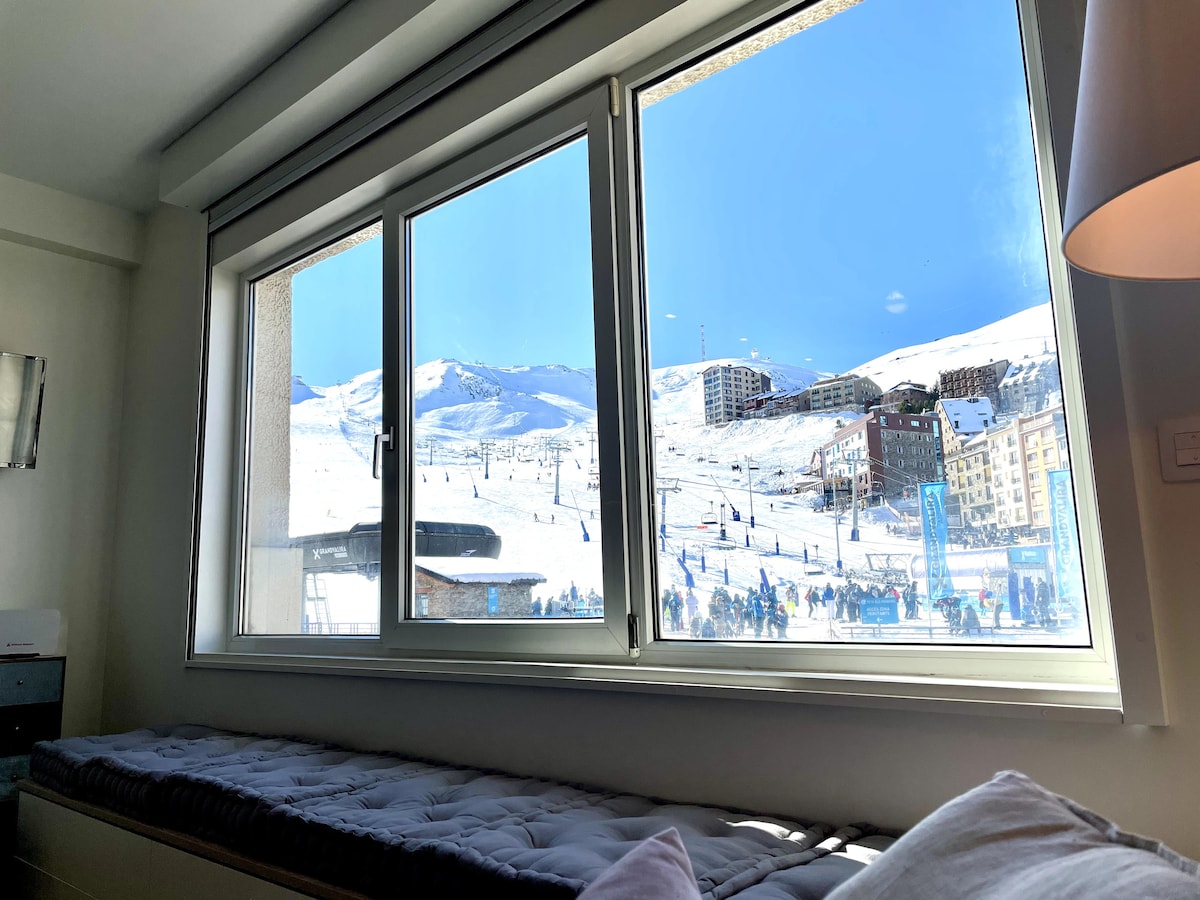 A large window offers a bright view of snowy ski slopes and distant mountains. Below the window, a cozy seat is visible, emphasizing the inviting atmosphere of the room. Natural light floods the space, enhancing the connection with the outdoor scenery.