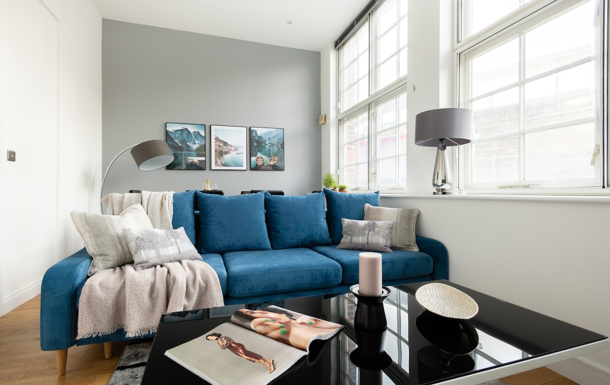A contemporary living area features a plush blue sofa adorned with soft cushions and a cozy throw. A sleek black coffee table showcases an art magazine and decorative elements. Large windows allow natural light to illuminate the room, complemented by stylish lamps.