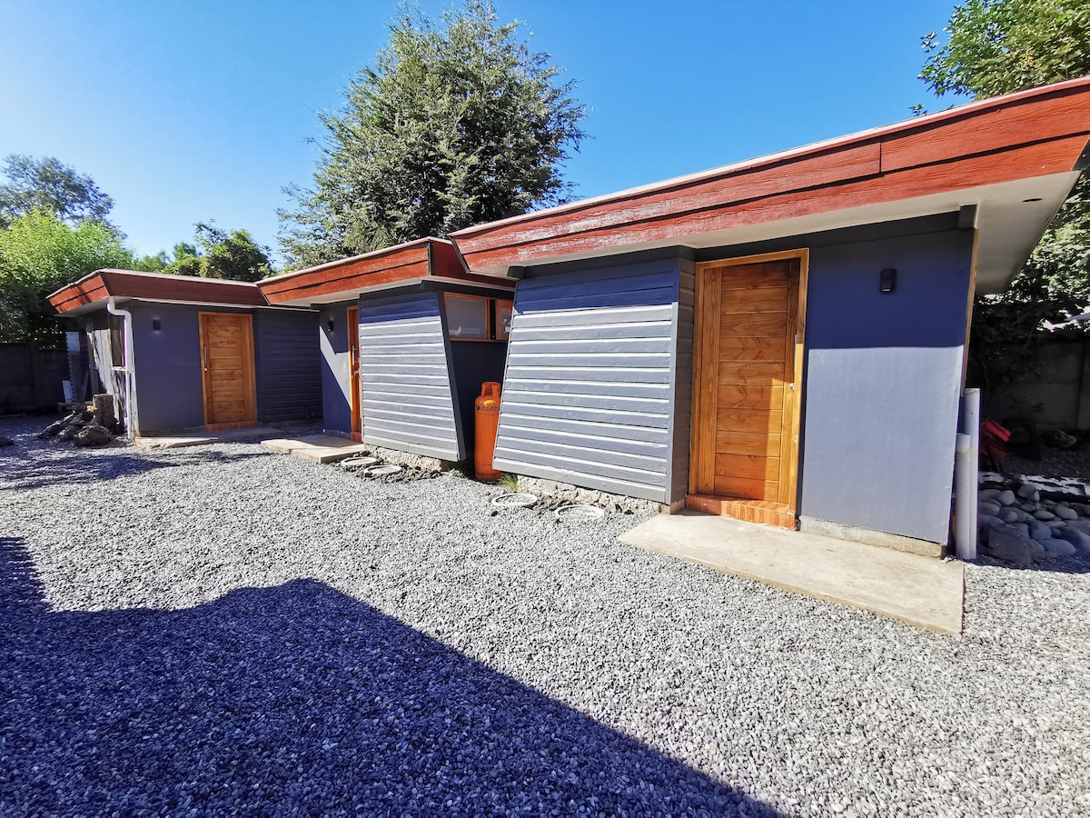 Two compact cabins are positioned side by side, featuring wooden doors and grey exterior walls. A pebbled pathway leads to each entrance, surrounded by greenery and clear blue skies, creating a peaceful outdoor setting.