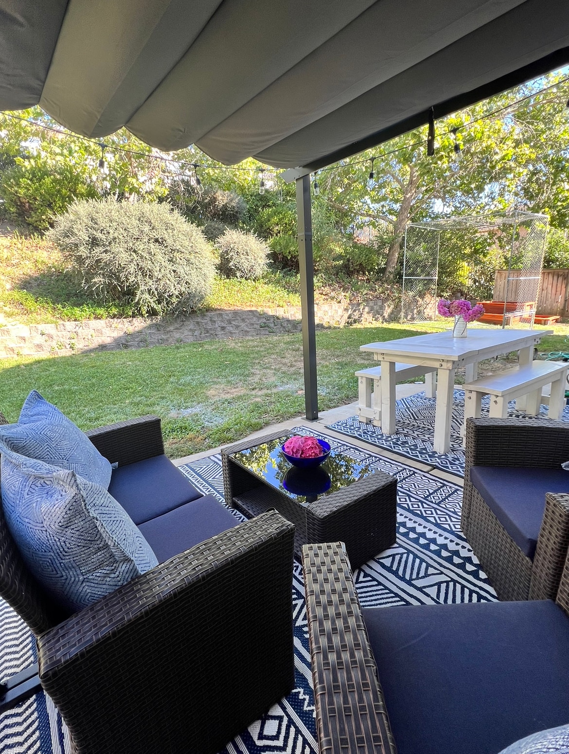 A covered outdoor seating area is showcased, featuring dark wicker chairs and a glass-top coffee table. A white dining table is positioned nearby on a patterned rug. Lush greenery and a garden are visible in the background, creating a refreshing outdoor space.