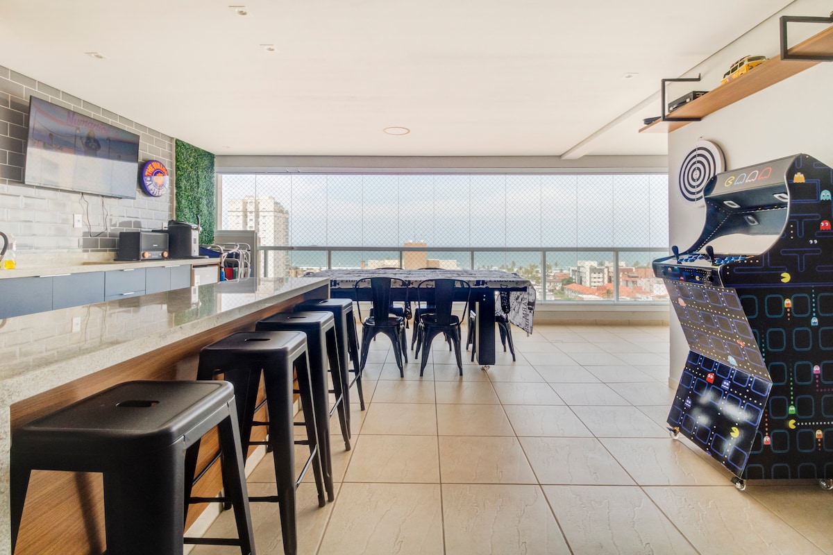 The gourmet balcony area features a long bar with several stools, a dining table designed for group meals, and a multi-game arcade machine. Large windows provide a view of the sea, allowing ample natural light to fill the space.