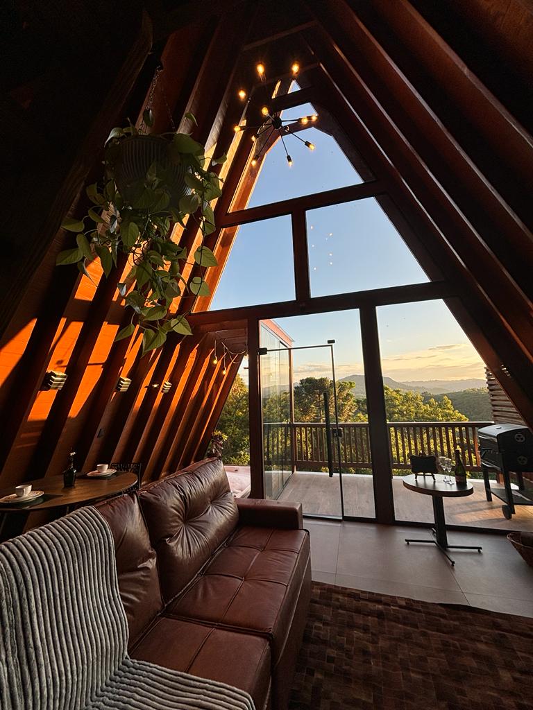 A cozy living area features tall, angled wooden walls with large glass doors that open to a scenic view. A brown leather sofa, adorned with a gray striped blanket, is complemented by a side table. Soft lighting and greenery enhance the inviting atmosphere.