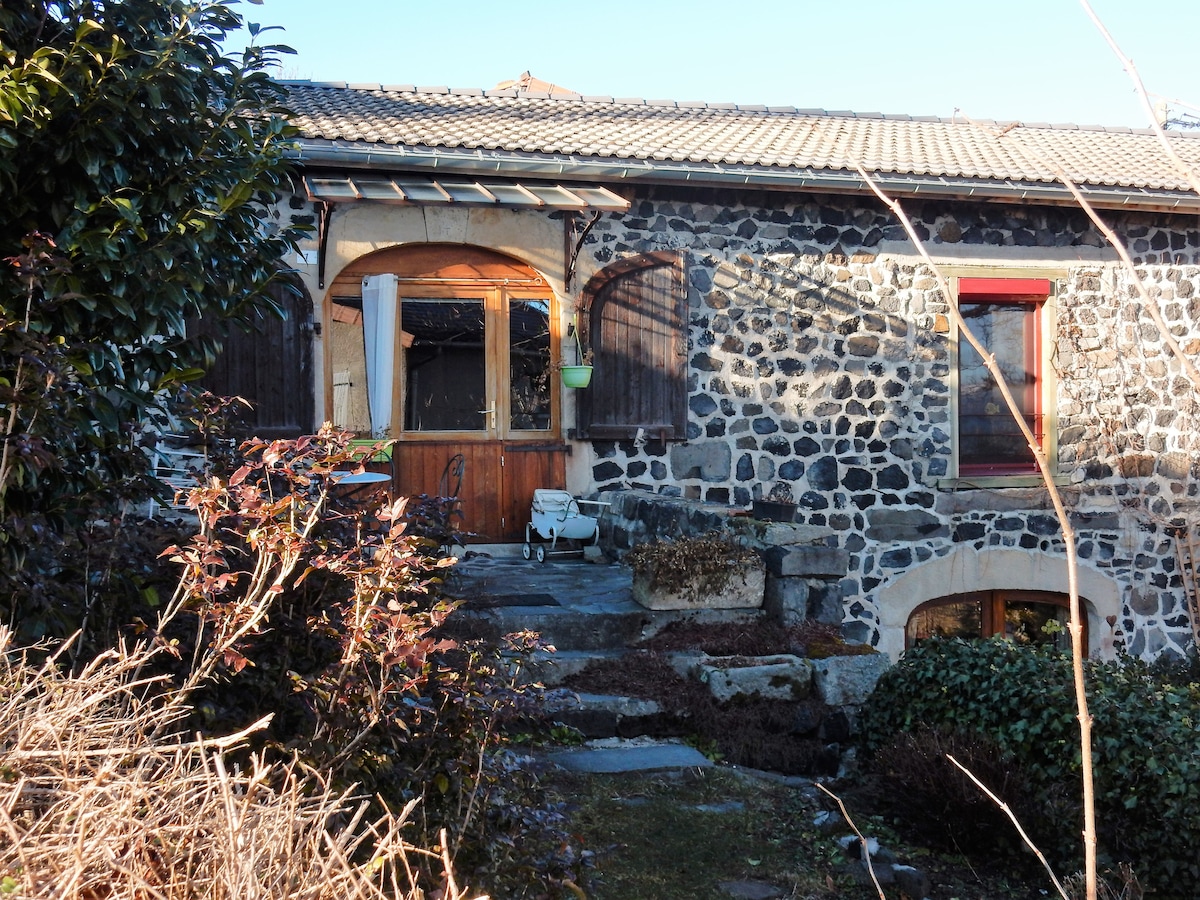 The exterior of a rustic stone building is framed by green foliage and gravel pathways. Wooden doors, accented with a small potted plant, offer a welcoming entry. Natural light highlights the textures of the stone, creating an inviting atmosphere in the countryside.