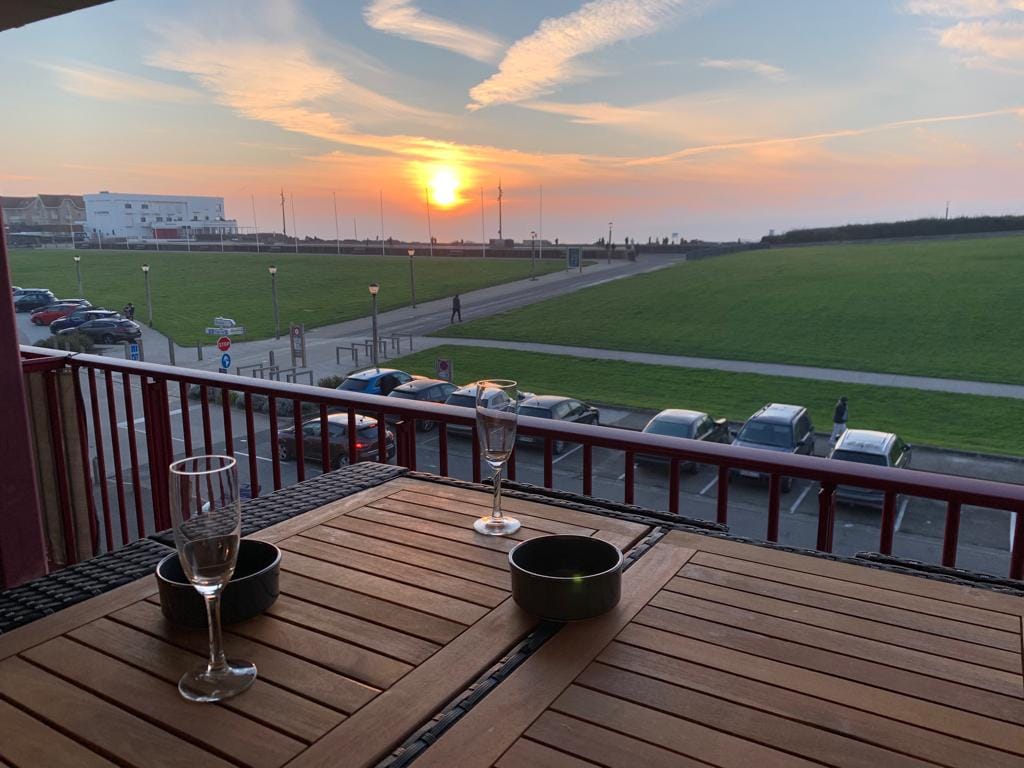 A table is set with two glasses and bowls on a wooden terrace, overlooking a grassy area. A sunset casts warm hues across the sky, while a line of parked cars is visible nearby. The scene captures a peaceful outdoor ambiance.