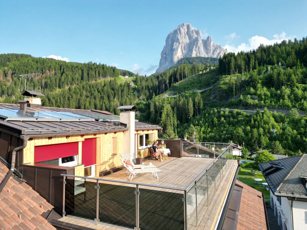 A spacious terrace is showcased with wooden decking and glass railings, providing stunning views of surrounding mountains and lush greenery. Sun loungers are positioned for relaxation, with a serene atmosphere illustrated by the clear blue sky and natural landscape.