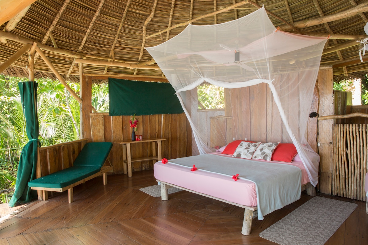 An open-air bungalow features a spacious bed draped with a protective mosquito net, complemented by soft bedding and decorative cushions. A lounge chair and a small table are situated beside the bed, with lush greenery visible through the wooden walls.