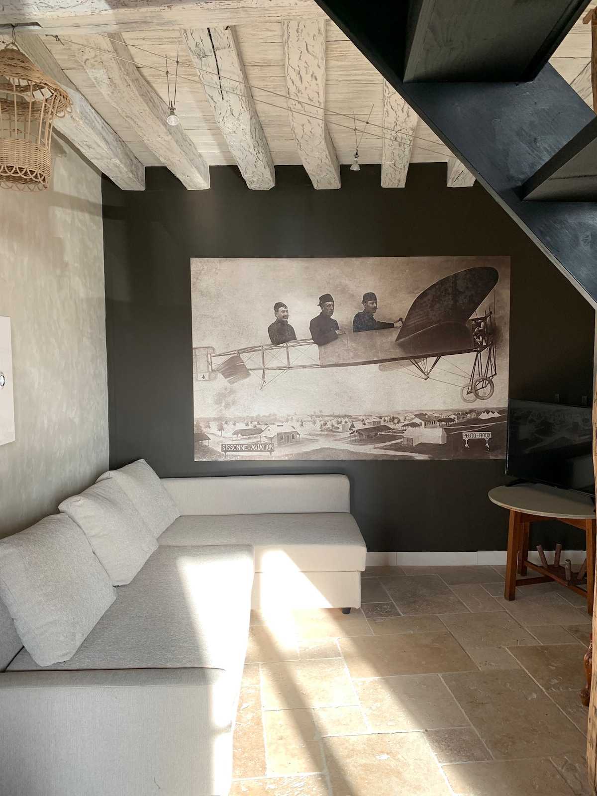 A cozy living space is depicted, featuring a light-colored sofa against a dark wall. An artistic black-and-white mural hangs above, depicting figures in an early aircraft. Natural light filters in, enhancing the room's inviting ambiance. A wooden table is positioned nearby.