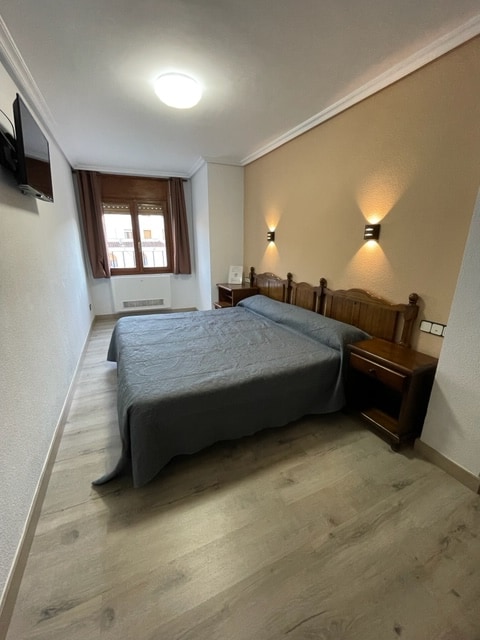 A cozy bedroom features a spacious bed with a dark grey blanket, flanked by wooden nightstands. Soft lights are mounted on the walls, providing gentle illumination. A window with light drapes allows natural light to enter, highlighting the room's neutral tones and wooden floor.