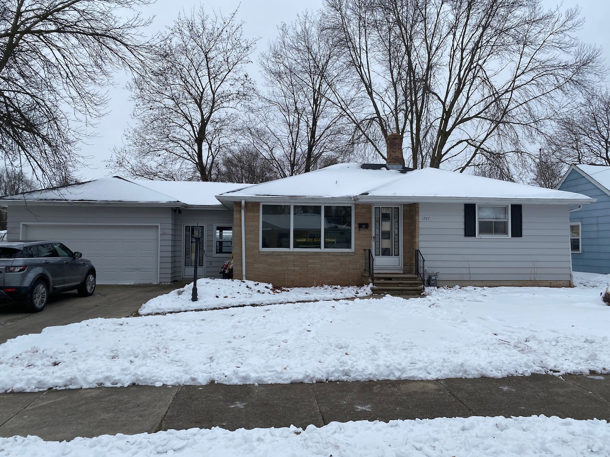 A single-story house features a light-colored exterior with a brick accent. The front porch has steps leading to the entrance. A driveway is visible, accommodating vehicles. Snow covers the ground, creating a serene winter landscape. Leafless trees are positioned on either side of the house.