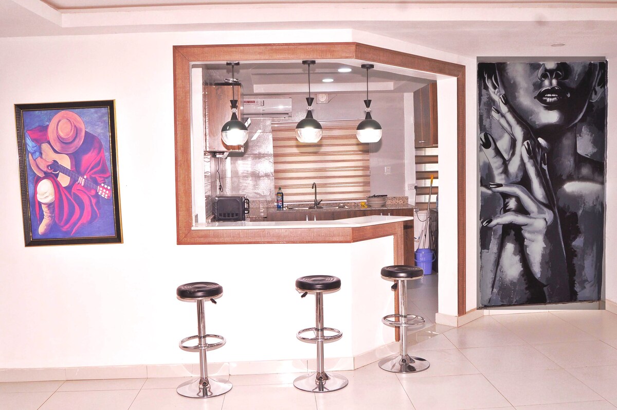 A modern kitchen area features a curved bar with three sleek stools positioned in front. Dark pendant lights hang above, illuminating the space. A bold artwork of a guitarist is displayed on one wall, while a striking mural of a hand is showcased on another.