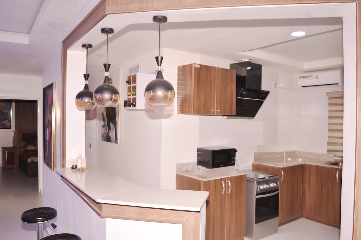 A modern kitchen is displayed, featuring wooden cabinetry and a granite countertop. Pendant lights hang above a bar area with stools. Essential appliances, including a microwave and stove, are visible, while the cooking space is brightly lit, contributing to a welcoming atmosphere.