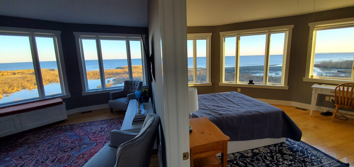 The spacious main bedroom features large windows offering panoramic views of the bay, filling the room with natural light. A comfortable seating area is visible beside a plush gray chair, and a cozy bed with a soft blanket is positioned toward the water view.
