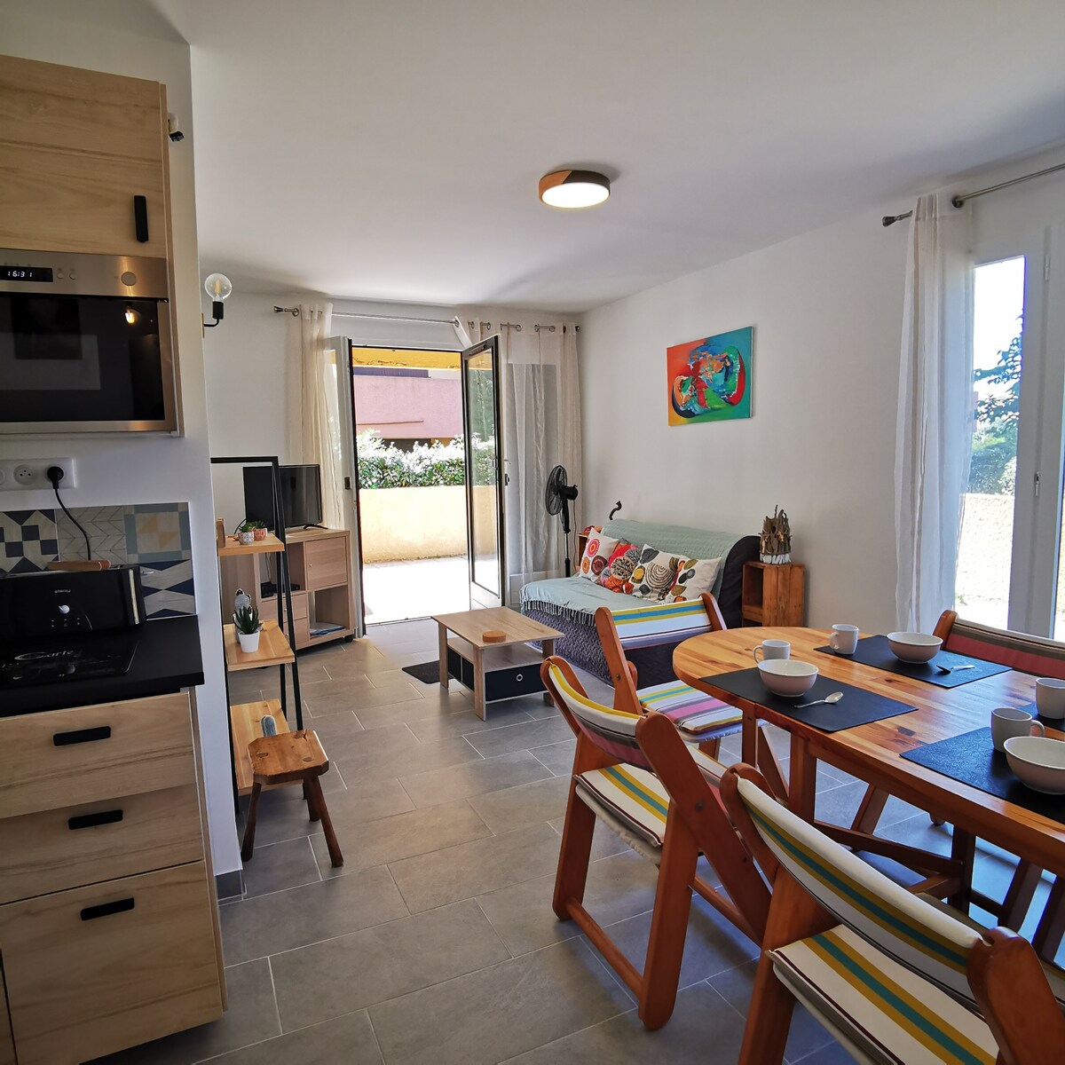A spacious living area features a comfortable seating arrangement and a dining table set for meals. Large glass doors lead to an outdoor terrace, allowing natural light to illuminate the room. Neutral tones and wooden accents create a welcoming environment, complemented by decorative wall art.