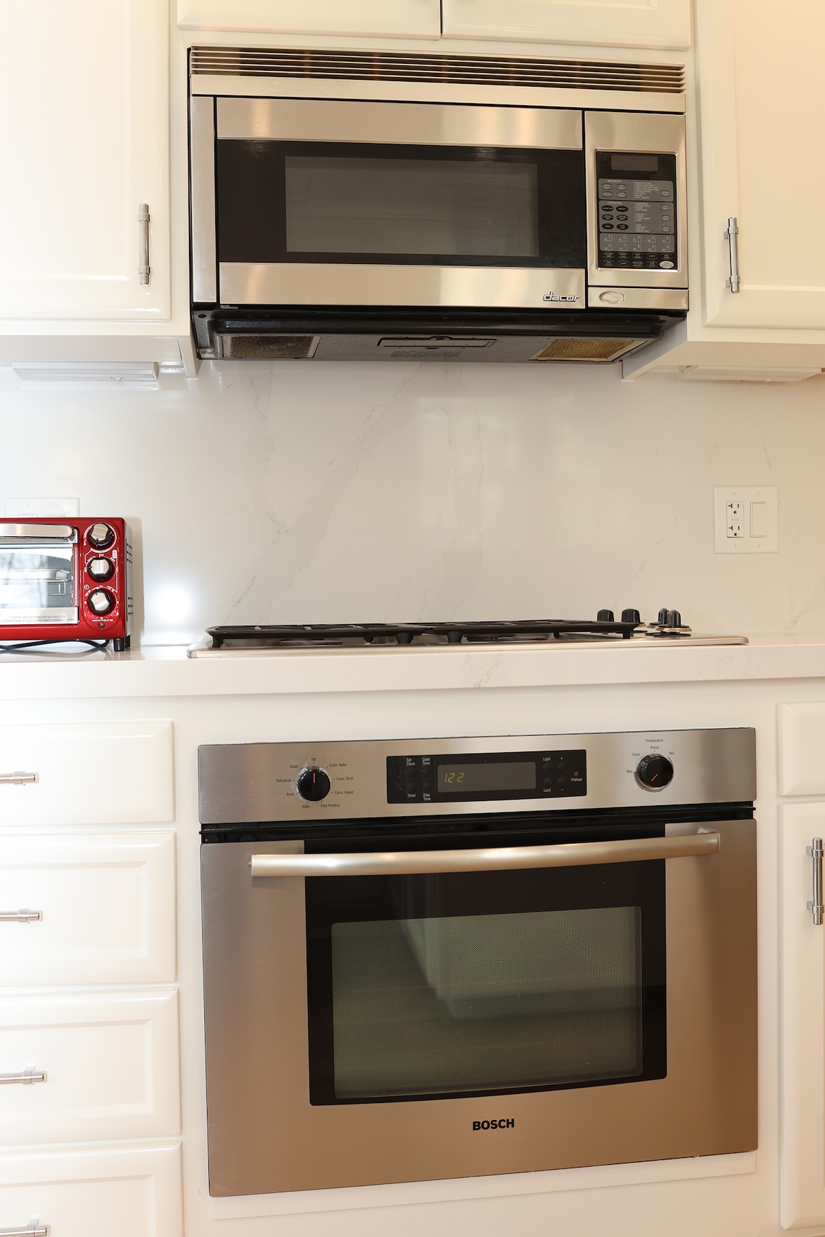 A sleek kitchen area is shown, featuring modern stainless steel appliances. A microwave is mounted above a gas stove, with a contemporary oven located beneath. White cabinetry and a light marble backsplash enhance the clean and functional design.