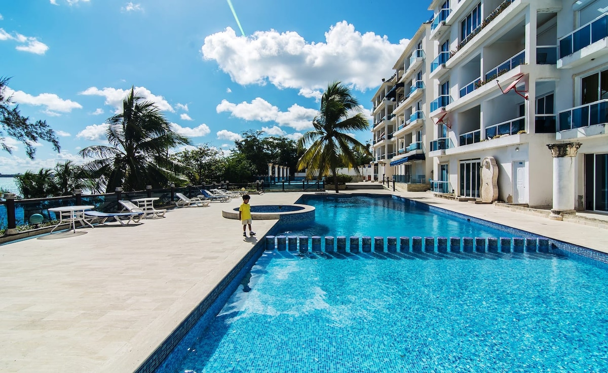 A large outdoor swimming pool features clear blue water, surrounded by lounge chairs and palm trees. Sunlight reflects off the water's surface, creating a bright atmosphere. The modern building stands in the background, with balconies overlooking the pool area.