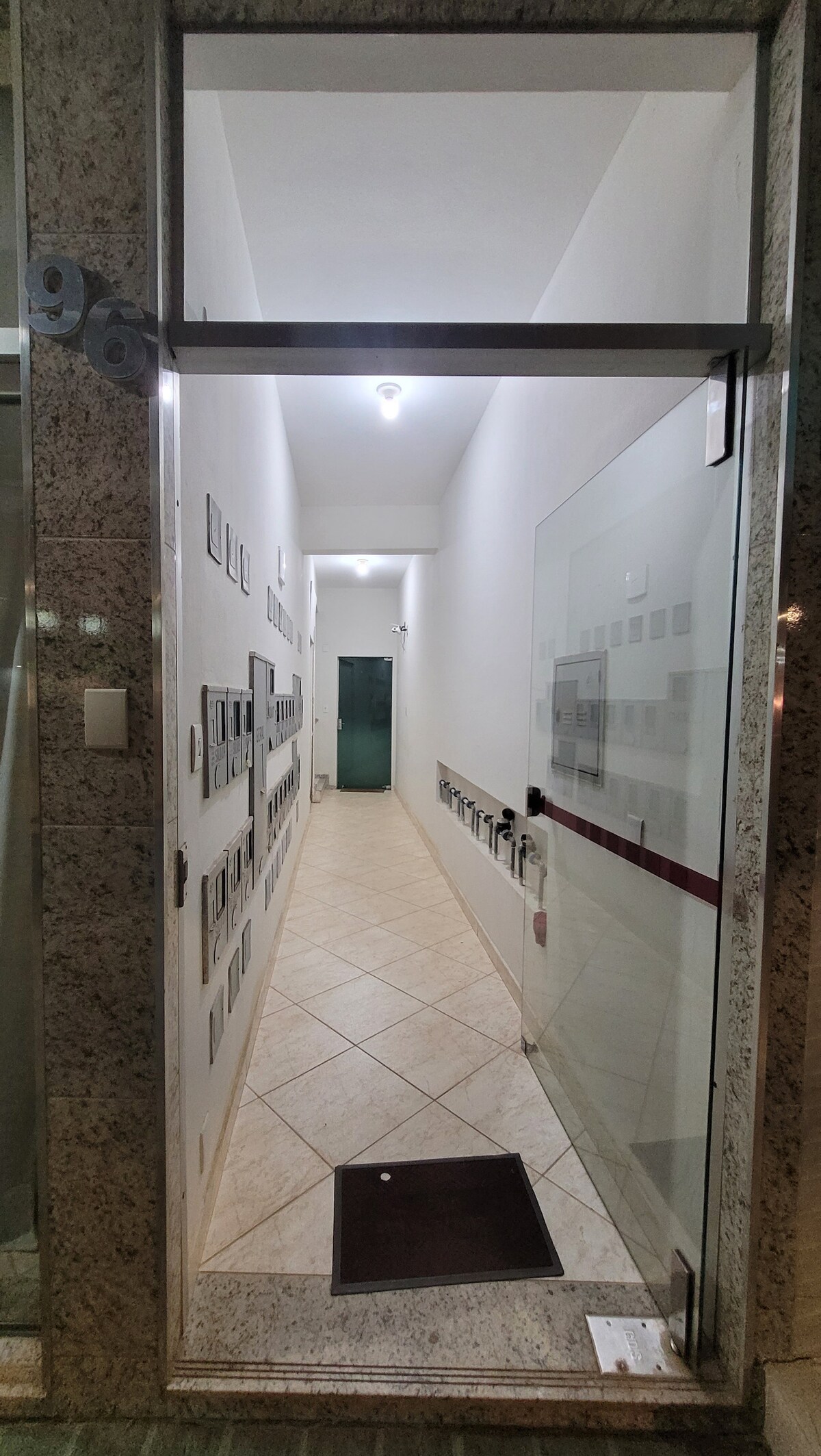 A well-lit corridor is shown, featuring smooth tile flooring and minimalist wall-mounted mailboxes on one side. A glass door leads to another area at the end of the hallway, which is framed by neutral-colored walls, creating a sense of openness and accessibility.