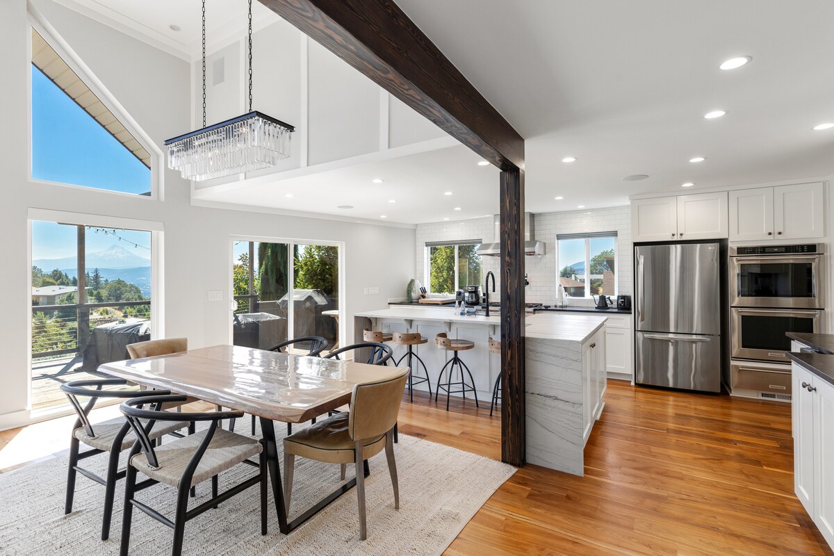 A modern kitchen and dining area are highlighted by large windows offering views of Mt. Hood. A spacious dining table with wooden chairs is positioned beside the kitchen, featuring contemporary appliances and an open layout. Natural light enhances the warmth of the wood flooring.