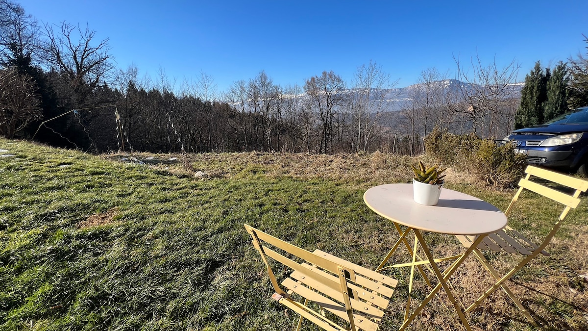 An outdoor seating area is depicted with a small round table and two folding chairs. A potted plant sits on the table. In the background, a landscape of trees and mountains is visible under a clear blue sky.