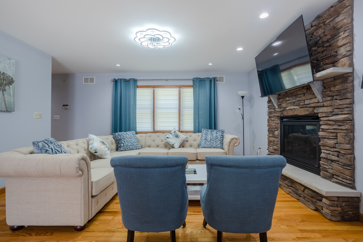 A well-lit living room features a comfortable tufted sectional sofa paired with two blue accent chairs. A stone fireplace adds texture to the room, while a mounted flat-screen TV offers entertainment. Decorative pillows and curtains in soft tones enhance the inviting atmosphere.
