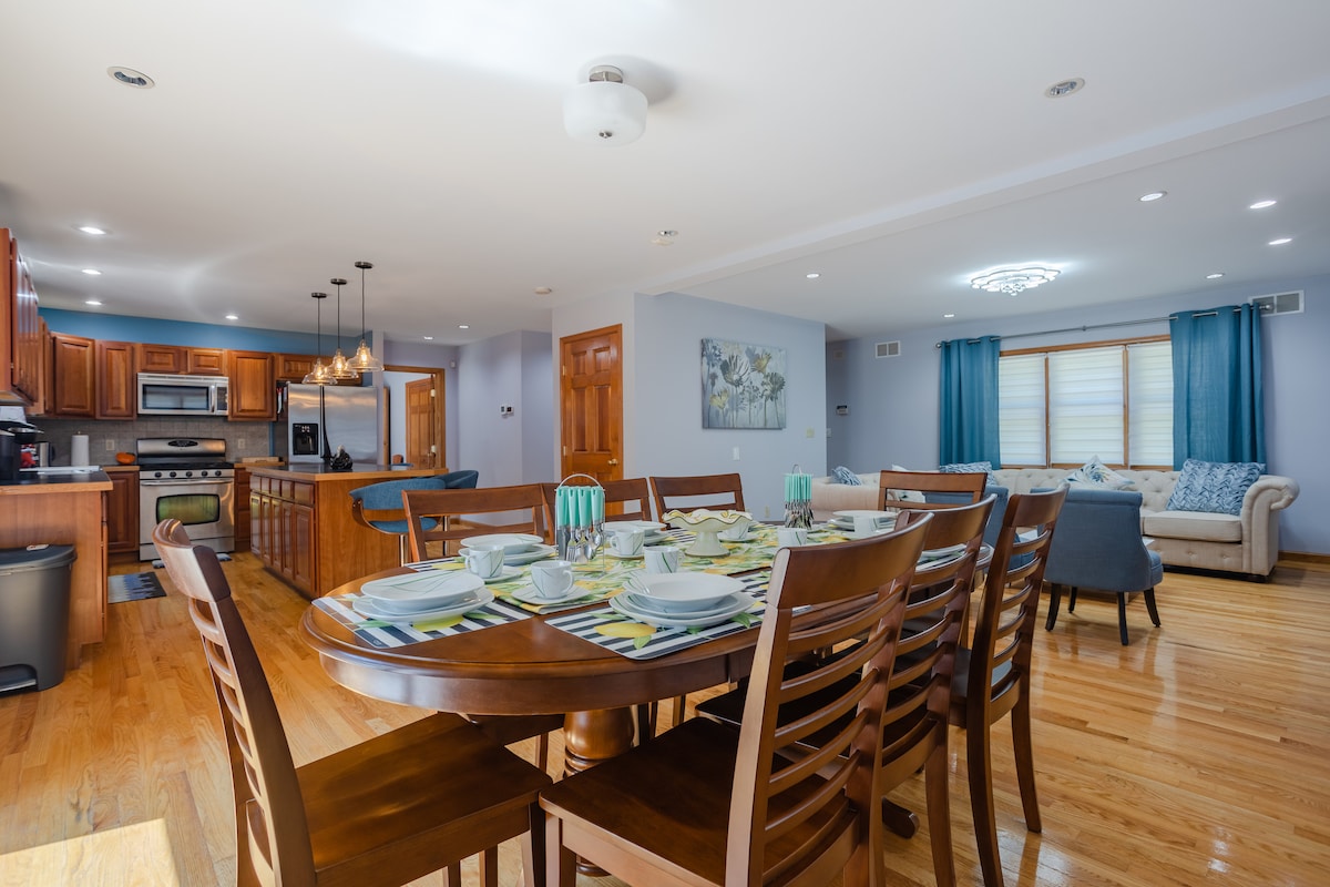 A spacious dining area is depicted with a large wooden table set for a meal, featuring plates and utensils. The open-concept layout transitions into a well-equipped kitchen with wooden cabinetry, alongside a cozy living room with comfortable seating and decorative accents.