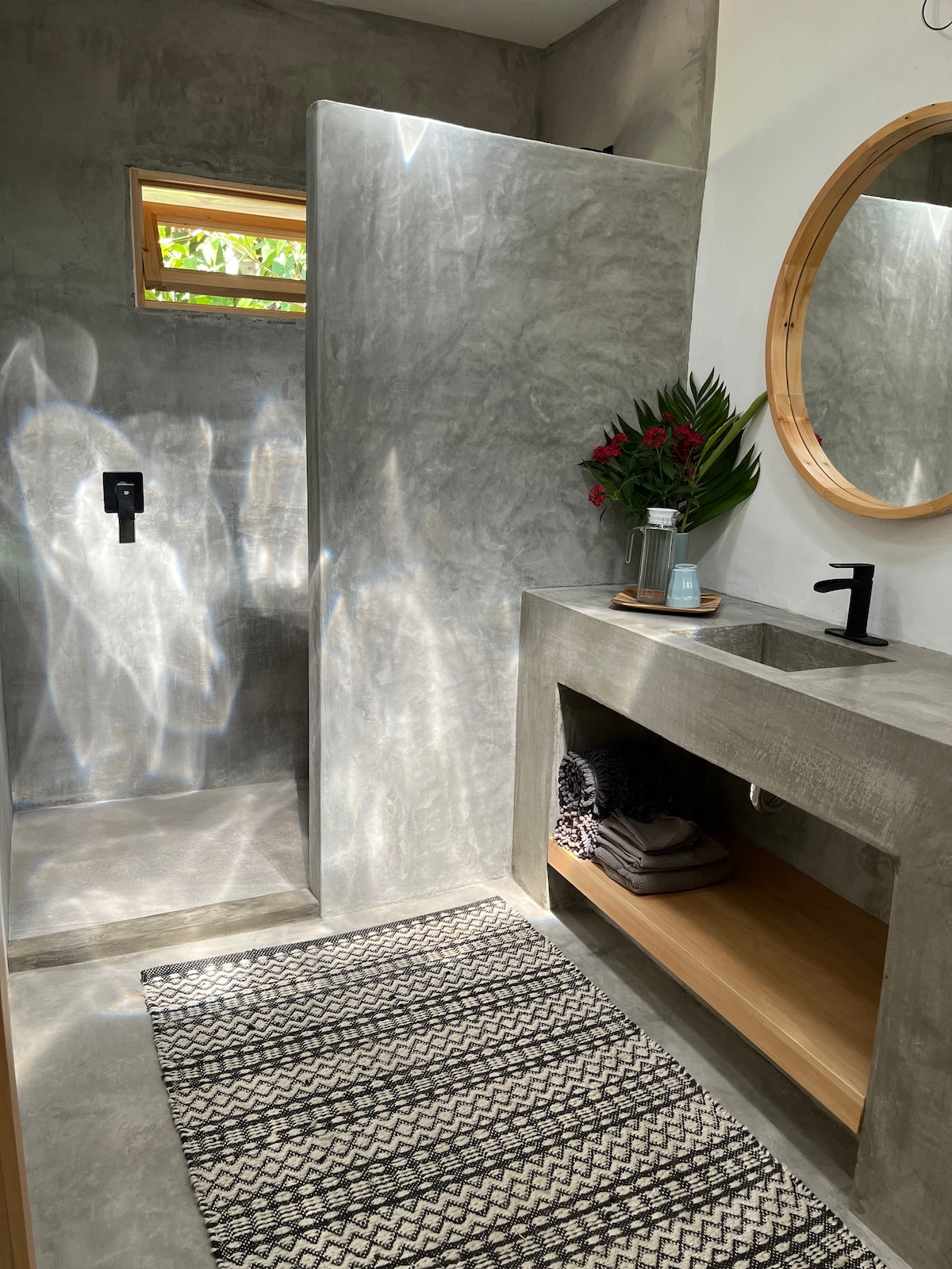 A modern bathroom features a walk-in shower with minimalist design and natural light filtering through a small window. A stylish cement sink is accompanied by a round mirror and wooden shelf displaying neatly arranged towels. A patterned rug adds warmth to the sleek, contemporary space.