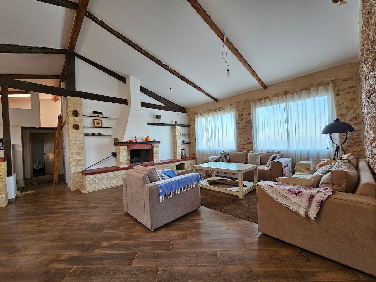A spacious living area features high, exposed wooden beams and large windows allowing ample natural light. Comfortable sofas are arranged around a central coffee table, and a stone fireplace provides a focal point. Light-colored walls and wooden flooring contribute to a warm ambiance.