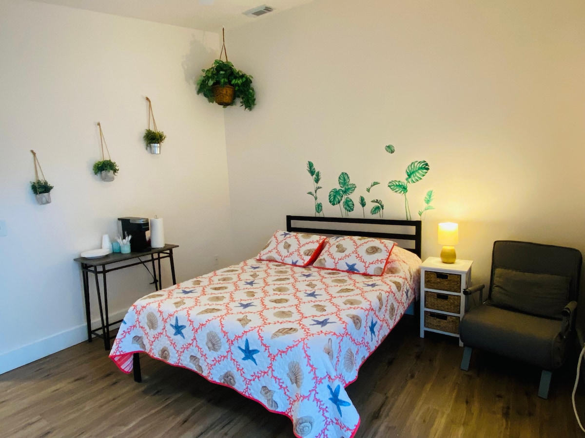 A comfortable bedroom features a queen size bed adorned with a patterned blanket. A small side table and chair are present, along with a coffee setup. Greenery decorates the walls, and a lamp provides soft lighting, creating a soothing environment.