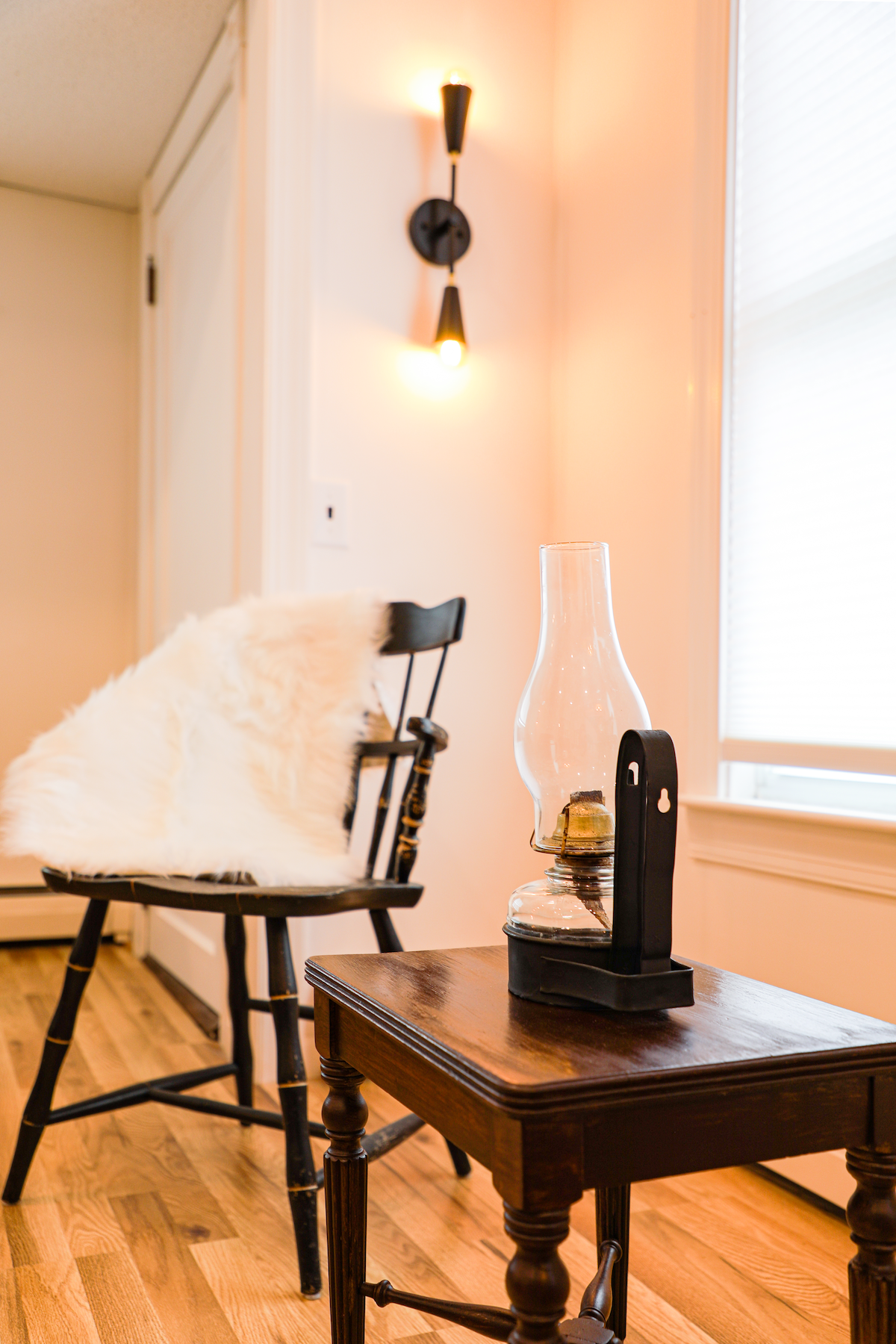 A wooden chair with a cozy white throw sits beside a small table. An oil lamp, gently glowing, rests on the table. Soft light from a wall sconce illuminates the space, enhancing the room's inviting ambiance.