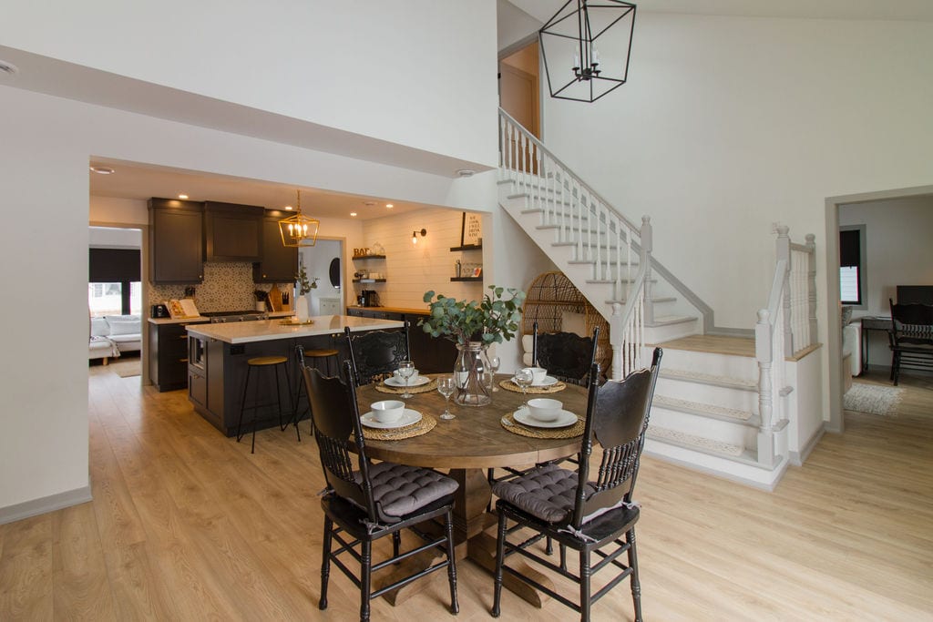 An open concept living space features a dining area with a round wooden table, surrounded by black chairs. The kitchen, equipped with modern appliances and dark cabinetry, is visible in the background, while a staircase with white railings leads to the upper level.