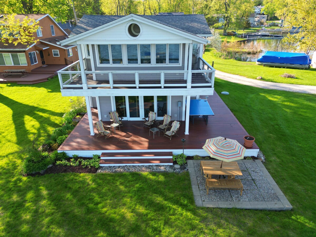 An aerial view captures a lakefront home surrounded by a spacious green yard. A large deck is furnished with outdoor seating, and an umbrella provides shade over a patio table. Nearby, a private dock and boats are visible in the serene waterfront.