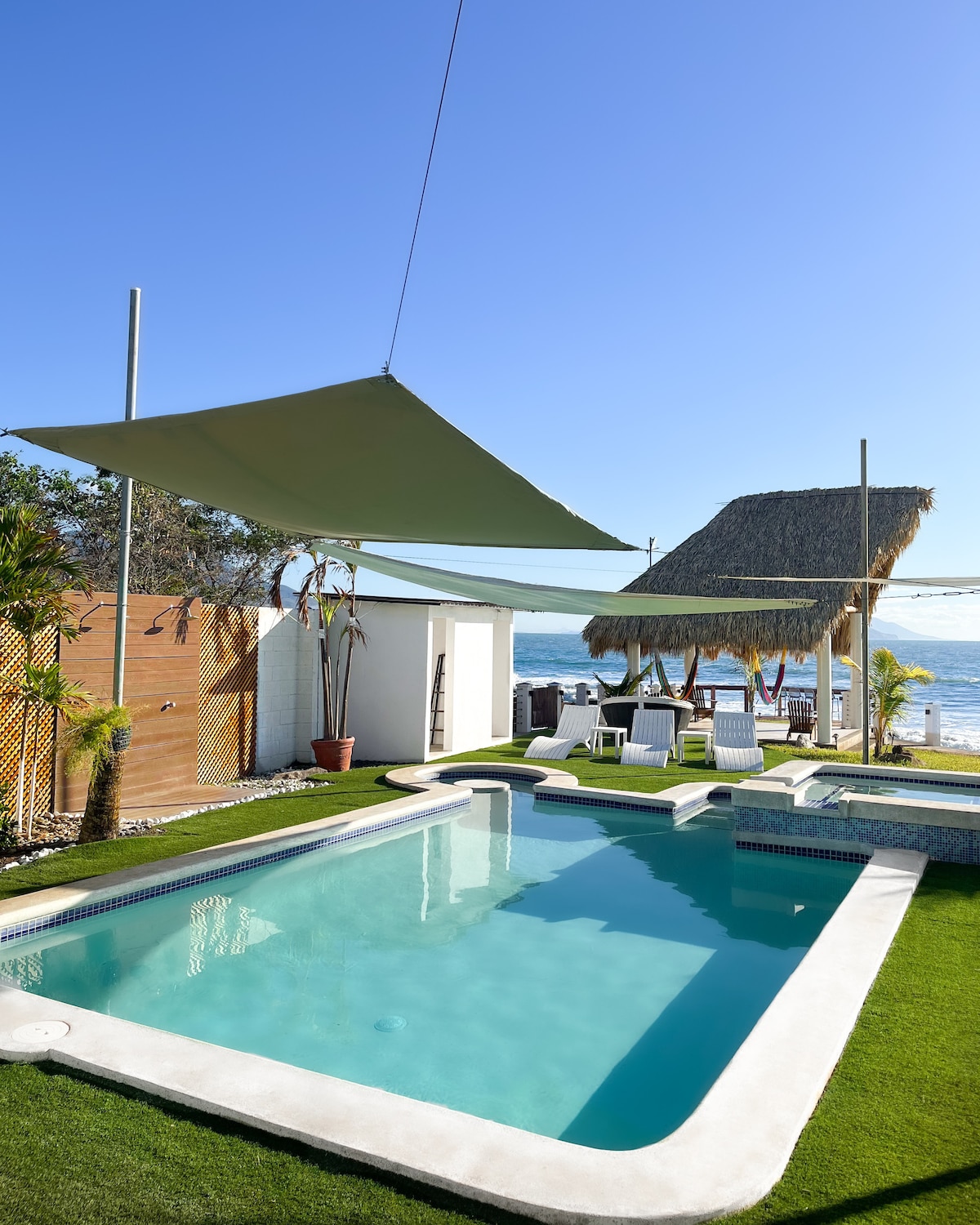 A serene outdoor area features a refreshing pool surrounded by green grass and beach chairs under shaded canopies. Thatched-roof gazebos provide additional seating, creating a relaxed environment with ocean views in the background.