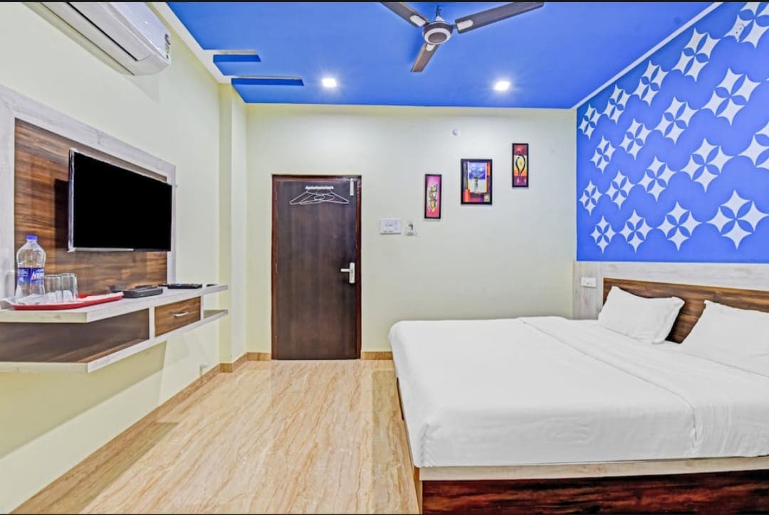 A spacious room features a plain wall adorned with decorative elements in blue and white. A large bed is positioned against the wall, complemented by a wooden headboard. A wall-mounted TV and a minimalist desk are also visible, along with a door leading to the hallway.