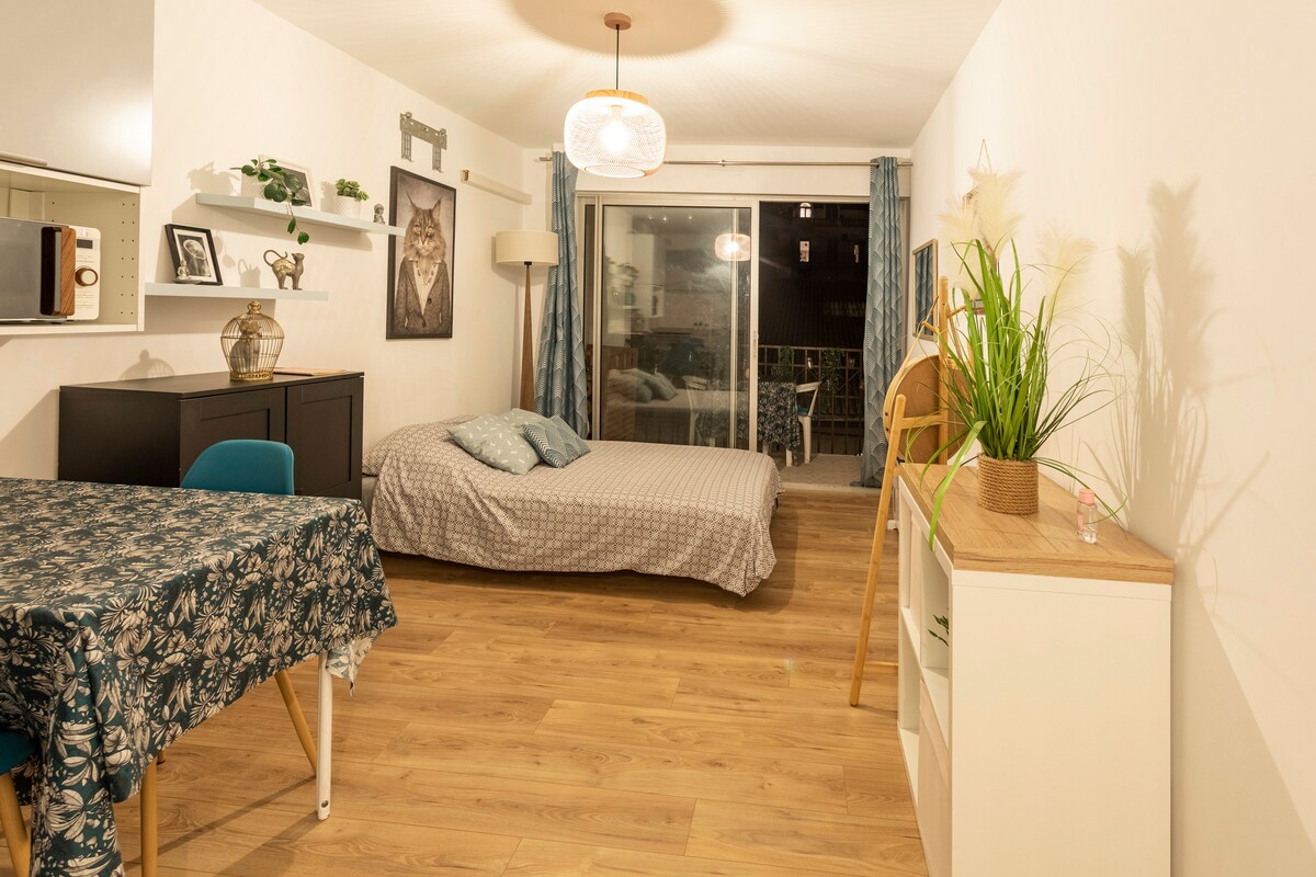 A bright room features a spacious bed with a patterned duvet and complementing walls. A dining table with blue chairs is positioned nearby, while a large glass door leads to an outdoor terrace. Decorative plants add a touch of greenery to the setting.