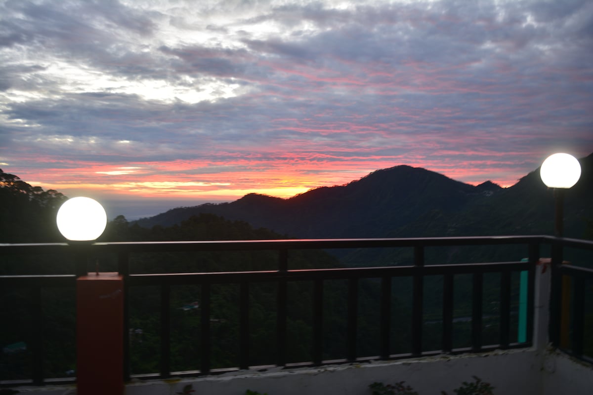 A private balcony offers stunning views of the mountains at sunset, with vibrant hues of orange and pink spreading across the sky. Two spherical lights illuminate the space, framed by a sturdy railing that enhances the outdoor setting.