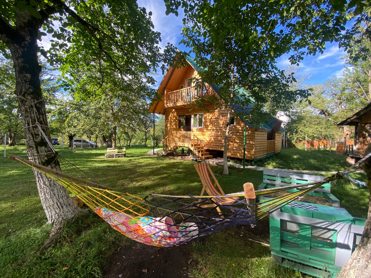 A hammock is suspended between two trees in a green garden, with a wooden cottage visible in the background. The cottage features a balcony and is surrounded by lush grass and trees, creating a peaceful outdoor space for relaxation.