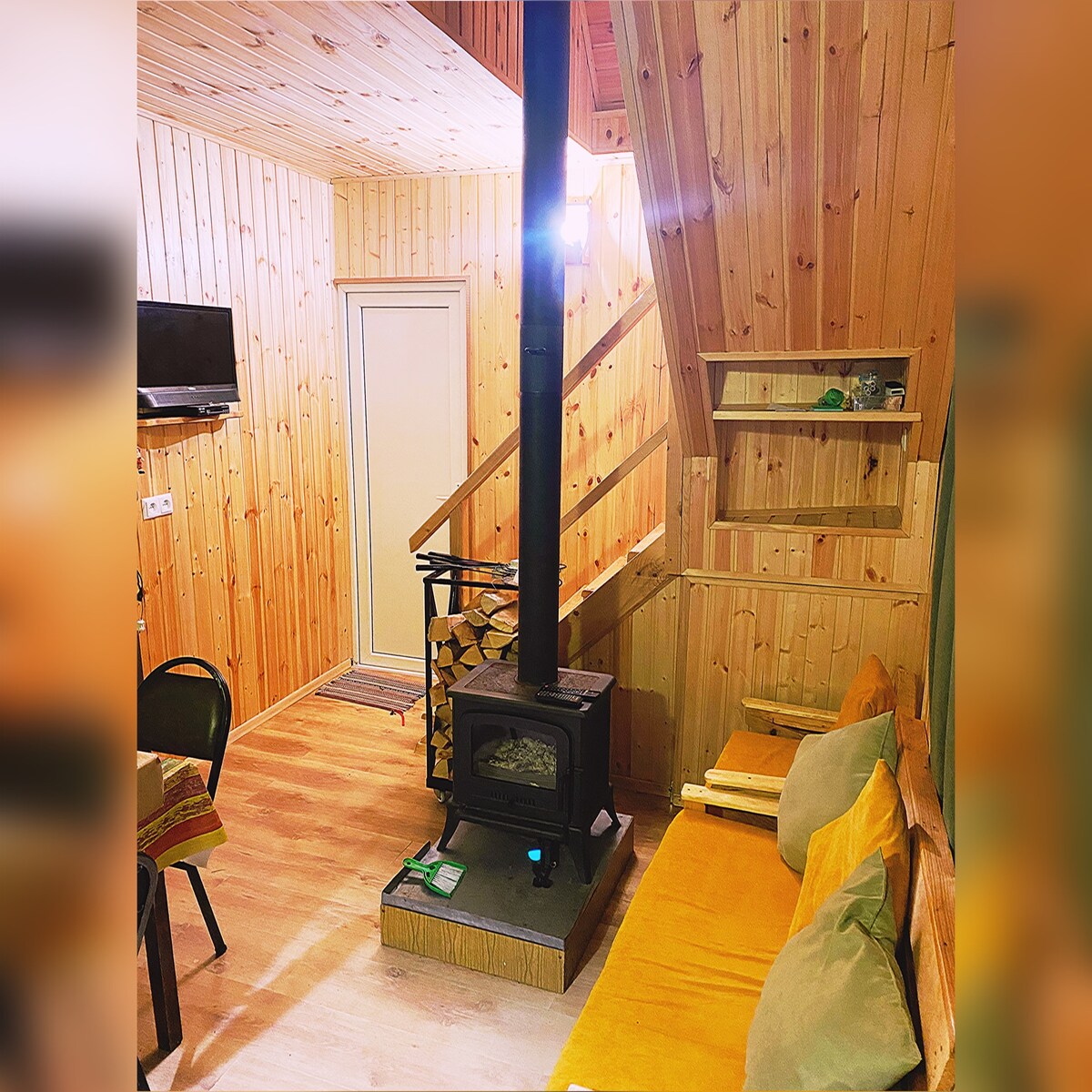 A wooden interior is showcased with a warm color palette. A compact seating area features a yellow sofa, while a freestanding stove is positioned on a wooden platform. A staircase leads to an upper level, complemented by shelving units. A wall-mounted TV is also visible.