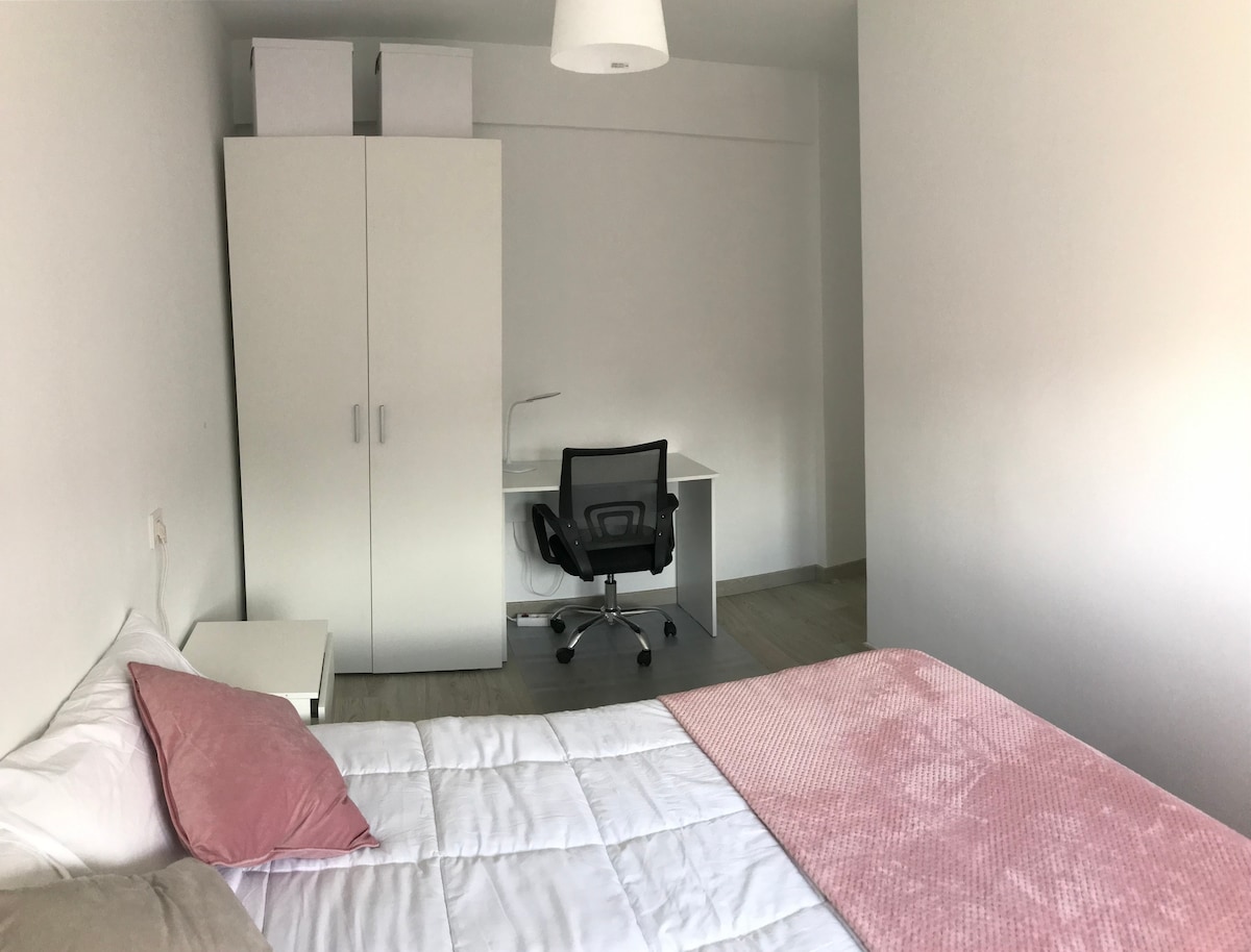 A bright bedroom is featured, showcasing a spacious bed dressed in white linens with a light pink throw. A desk and rolling chair are positioned adjacent to a tall white wardrobe, providing ample storage. Natural light fills the space, enhancing its airy feel.