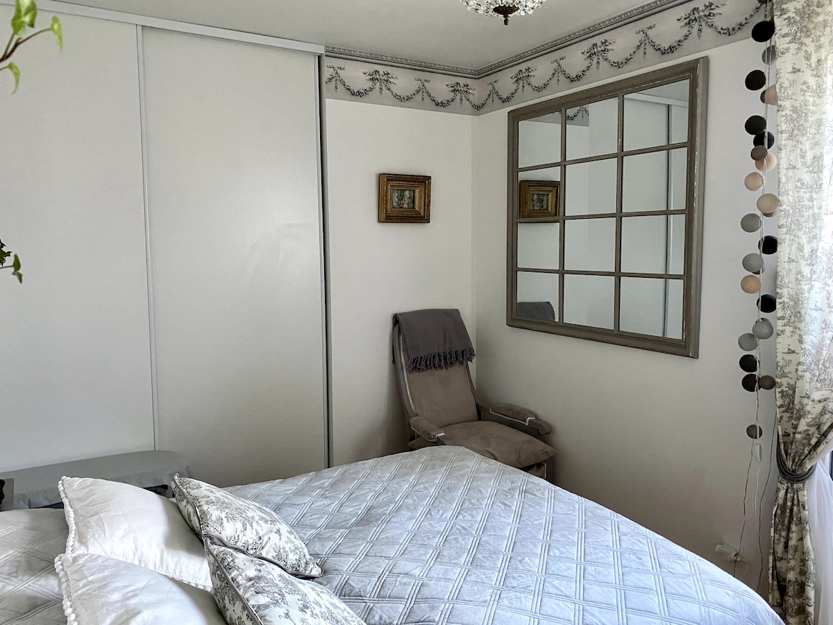 A cozy bedroom is showcased, featuring a spacious bed adorned with plush pillows. A mirror with a grid design reflects the soothing colors of the room. A comfortable chair sits nearby, accompanied by decorative elements including a small plant and draped curtains.