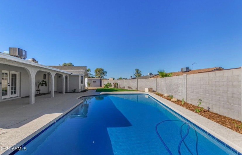 A tranquil outdoor space features a clear, blue pool surrounded by concrete decking. The area is enclosed by a low wall, and a small guest house is visible in the background. Lush plants line the perimeter, adding a touch of greenery.