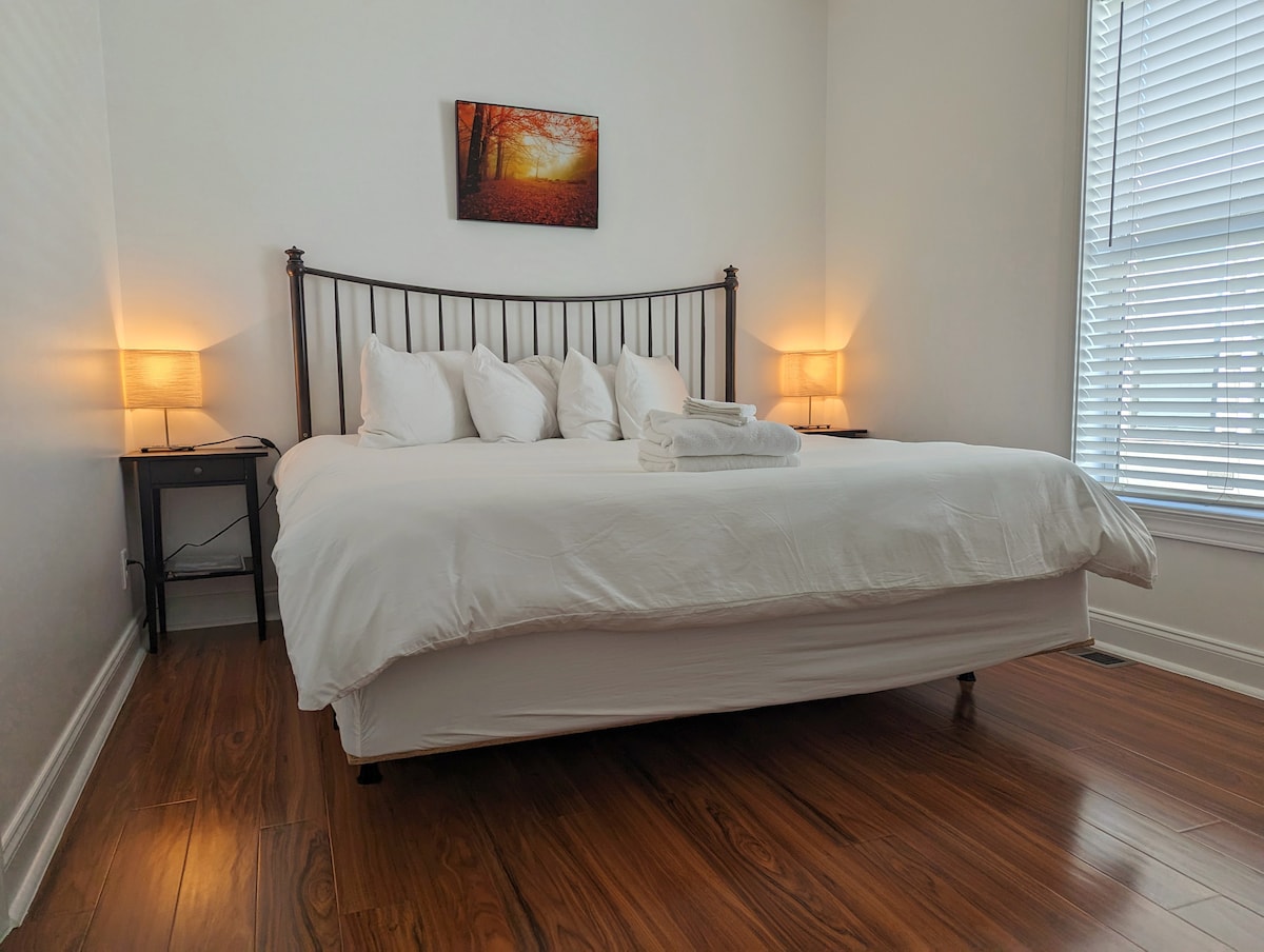 A spacious bedroom is furnished with a large bed adorned with crisp white linens and multiple pillows. Soft bedside lamps provide gentle illumination, while a piece of artwork depicting autumn leaves adds a warm touch to the wall. Natural light enters through a window with white blinds.