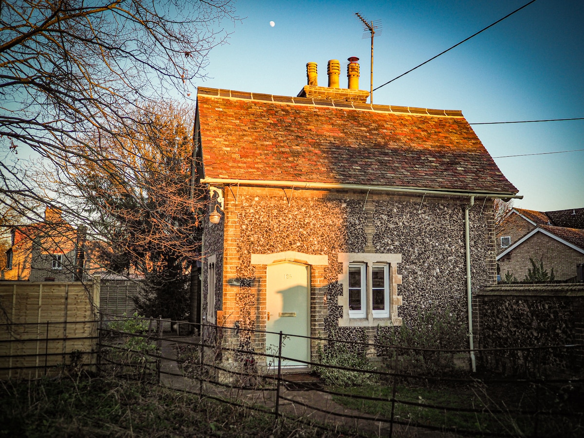 The historic gatehouse features a charming flint exterior with a red-tiled roof. A welcoming light green door stands out, surrounded by multiple windows. The scene is complemented by the soft glow of the setting sun and a glimpse of the moon in the sky.