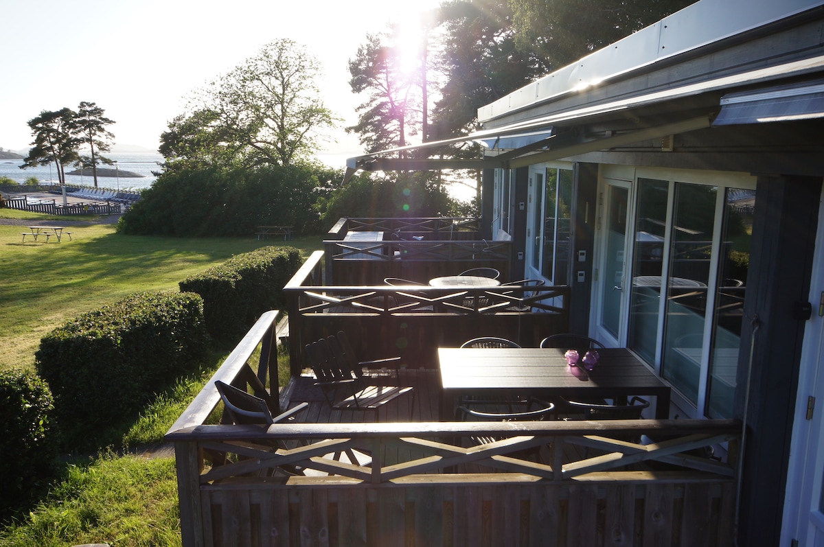 A wooden deck is visible, featuring outdoor dining furniture, including a table and chairs. Lush greenery surrounds the space, with distant views of the water and trees. Sunlight casts a warm glow, highlighting the inviting atmosphere of the outdoor seating area.