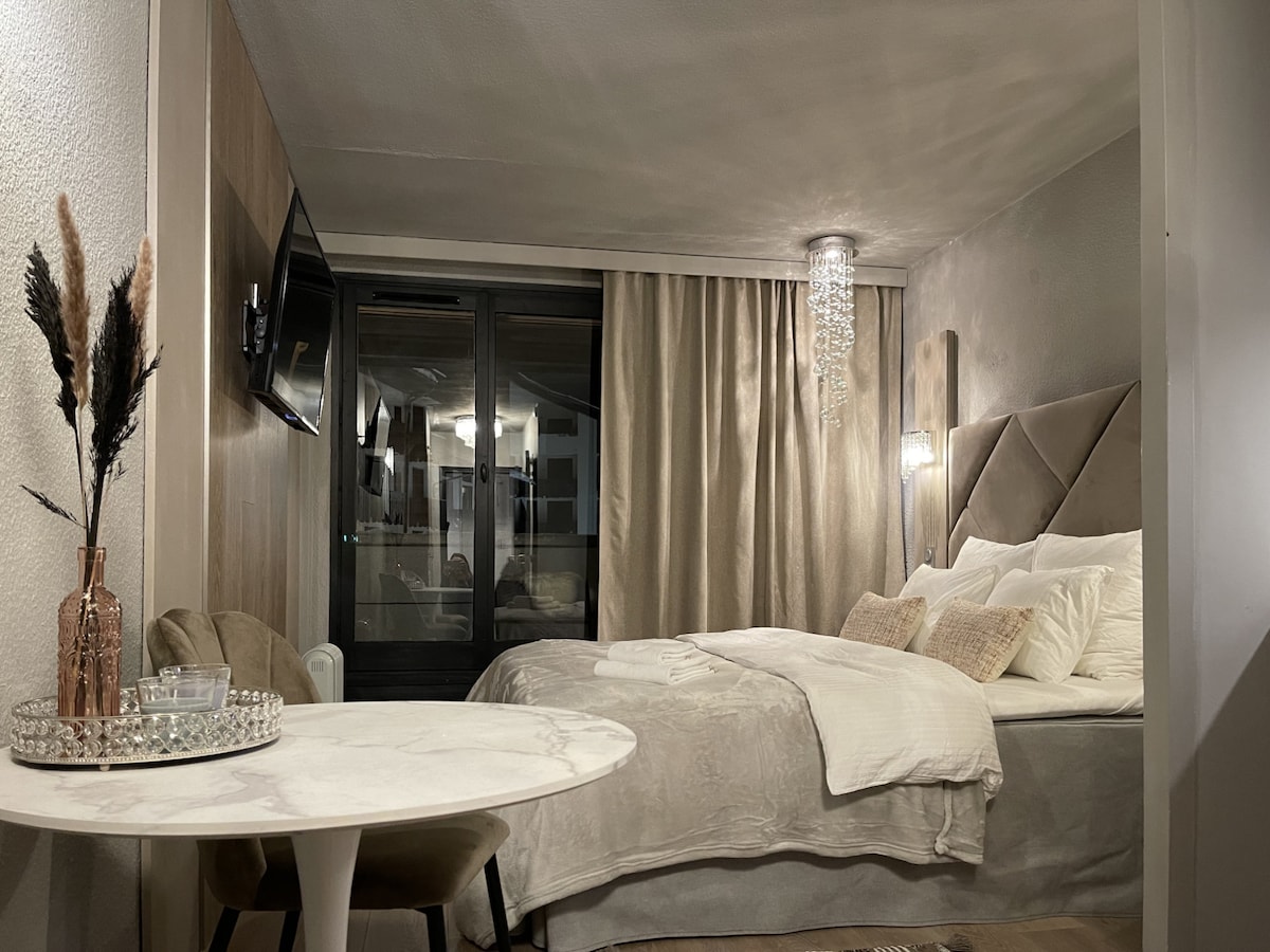 A modern and softly lit studio features a spacious bed with elegant linens and decorative pillows. A small round table sits beside plush seating, while a TV is mounted on the wall opposite. Large glass doors lead to a balcony, enhancing the open feel.