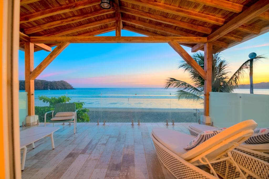A covered outdoor patio displays comfortable lounge chairs and a cozy seating area, with a stunning ocean view framed by palm trees. The sky transitions in vibrant hues, reflecting on the tranquil waters below.