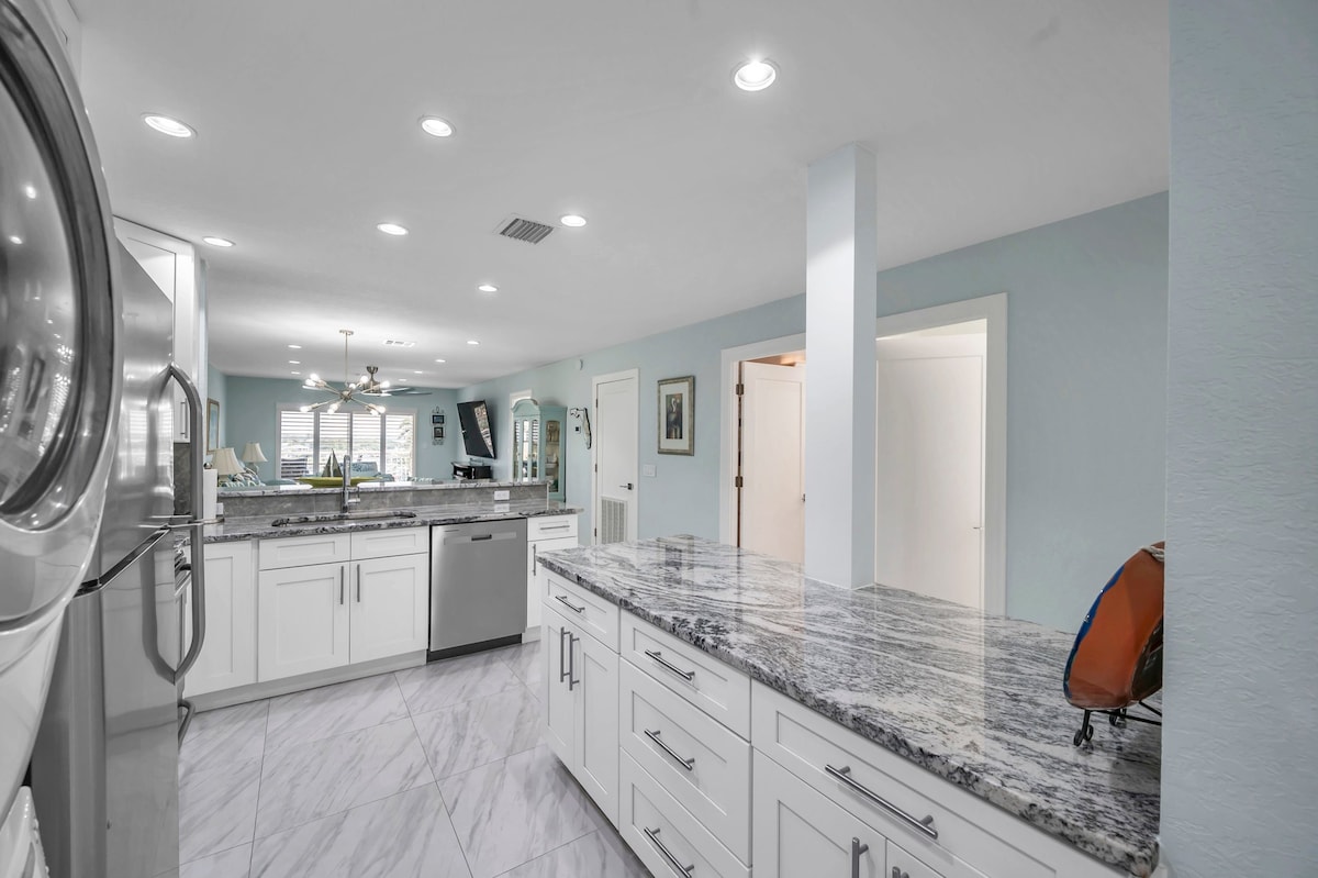 The spacious kitchen features modern appliances and granite countertops, offering ample workspace. A light color palette enhances the bright atmosphere, while multiple light fixtures illuminate the area. Access to the living space and adjacent rooms is visible through open doorways.