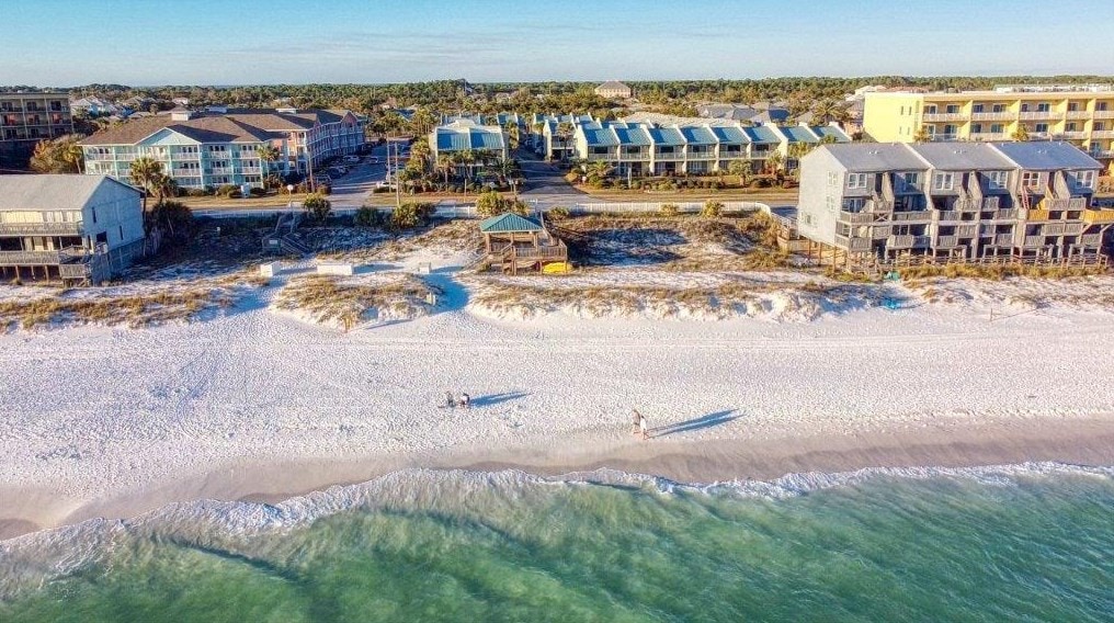 An aerial view captures the serene shoreline with soft white sands meeting clear turquoise waters. A nearby beach gazebo offers shade, while beachside properties line the coast, revealing a quiet community atmosphere. The landscape showcases a blend of natural beauty and coastal living.